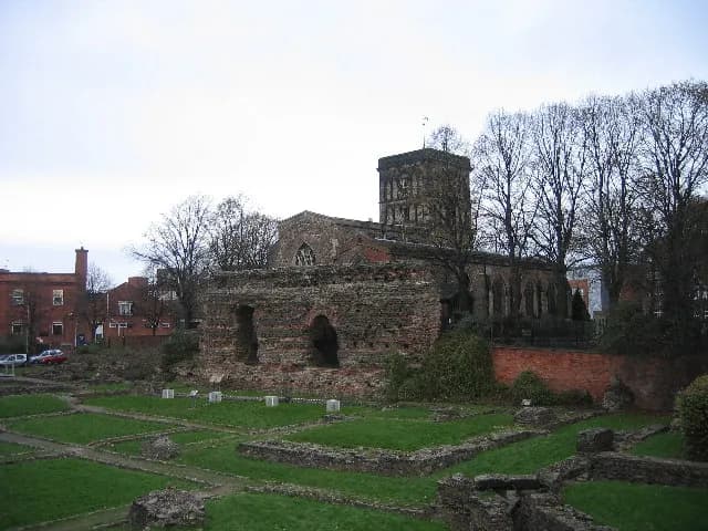 Ancient stone ruins and green lawns lie in the foreground of a large brick building with a tall square tower and bare trees.
