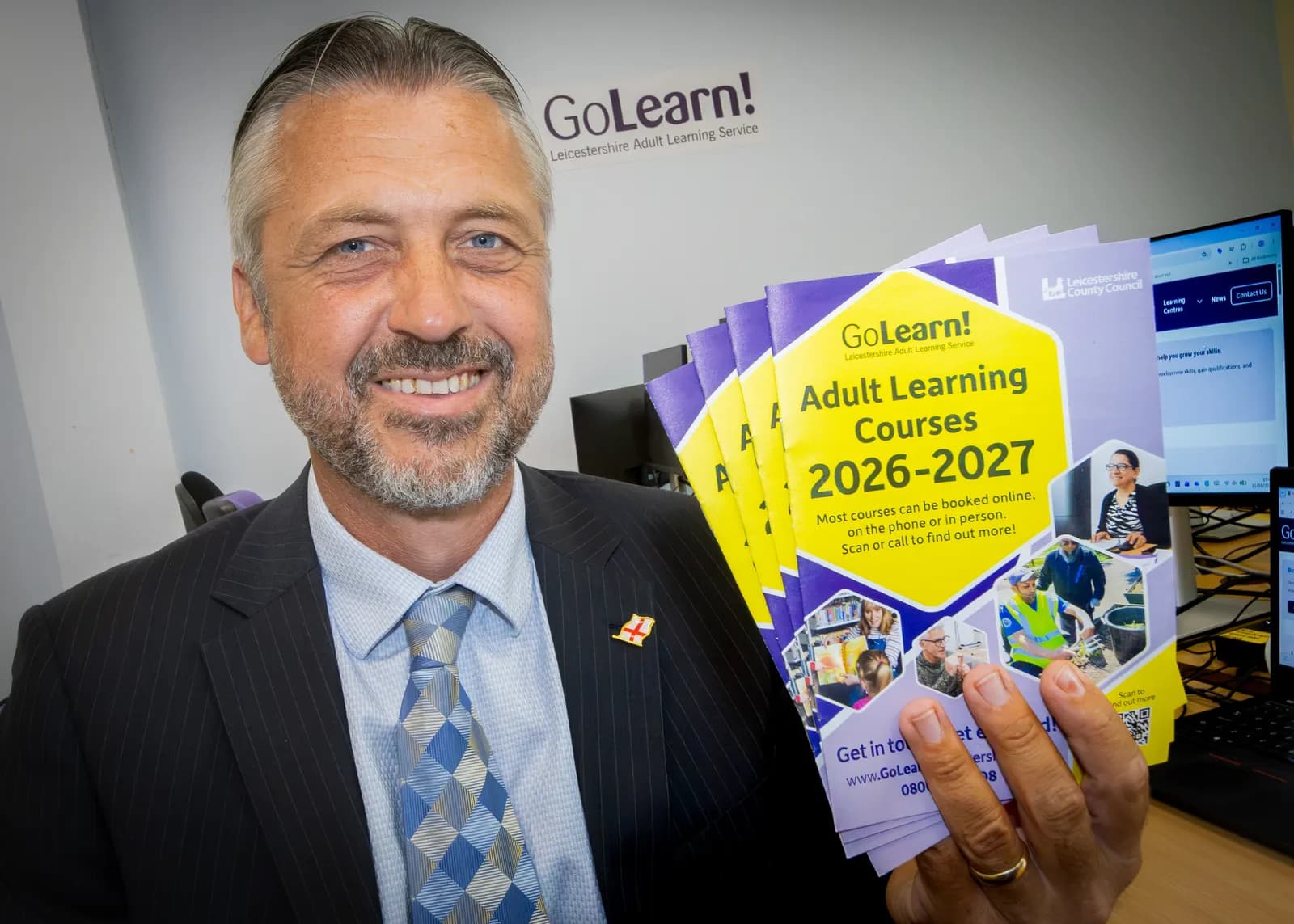 A smiling man with graying hair and a beard holds up a stack of 'Adult Learning Courses 2026-2027' brochures in an office setting.